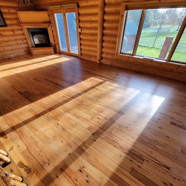 Finished hardwood floor in a log cabin great room with fireplace and a window view of a pasture, sunlit