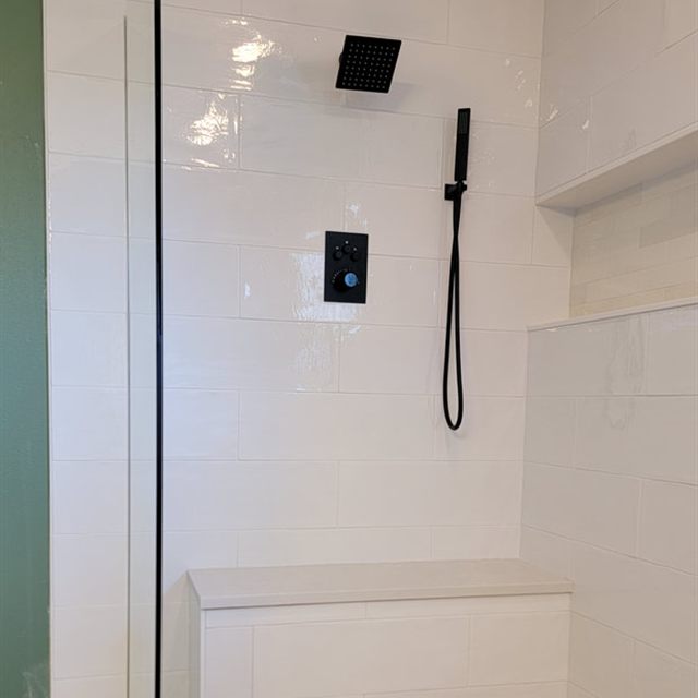 Wide view of finished white tile shower with black fixtures, niche, bench, and pebble floor