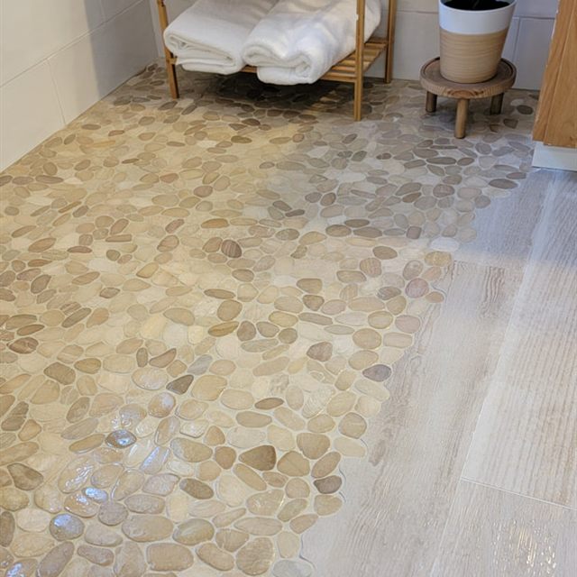 Transition from pebble mosaic shower floor to wood-look plank tile, with staged towels and a plant
